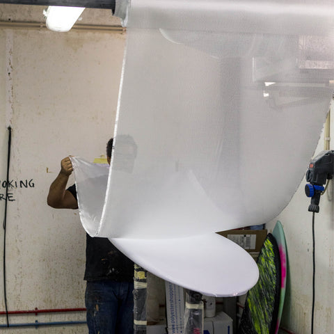 A man applying fiberglass cloth to a shaped surfboard blank.